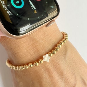 May include: A gold beaded bracelet with a white star charm. The bracelet is on a person's wrist.