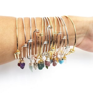 May include: A stack of gold, silver, and rose gold bangle bracelets with small, colorful gemstone charms hanging from each one.