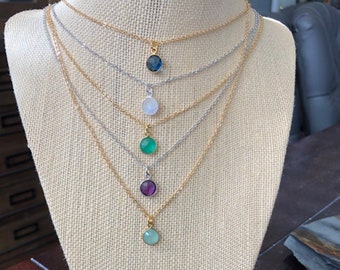 May include: Five gold necklaces with different colored gemstone pendants on a fabric mannequin. The pendants are blue, white, green, purple, and light green.