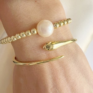 Könnte beinhalten: Ein goldfarbenes Perlenarmband mit einem großen weißen Perlenanhänger und ein goldfarbenes Schlangenarmband.