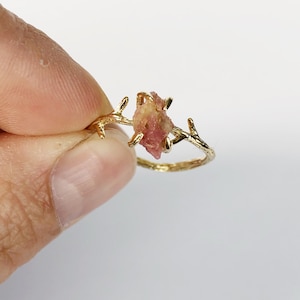 May include: A gold ring with a delicate branch design and a pink gemstone. The ring is set on a white background.