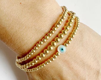 May include: Three gold beaded bracelets stacked on a wrist. One bracelet features a small, round charm with a blue eye design. The bracelets are made of small, round, gold-colored beads.