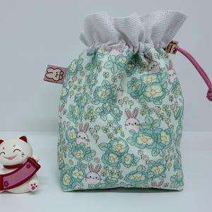 Puede incluir: Una pequeña bolsa con cordón con un estampado floral y de conejos en tonos verdes y azules. La bolsa tiene una parte superior de malla blanca y un cordón rosa. A la izquierda hay una figura de gato blanco y rojo.