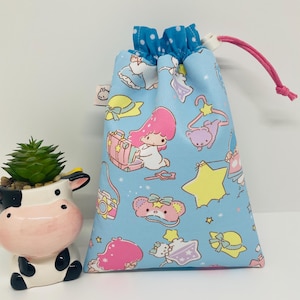 Può includere: Un sacco con coulisse azzurro con un motivo ripetuto di personaggi dei cartoni animati, stelle e accessori. La borsa ha una parte superiore a pois blu e un cordoncino rosa. A sinistra c'è una fioriera a forma di mucca in ceramica con una pianta succulenta.