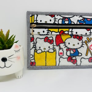 May include: A gray zippered pouch with a white background and a colorful print of Hello Kitty characters. The pouch has a gray border and a gold zipper.