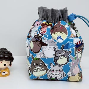 May include: A blue drawstring bag featuring a pattern of cartoon characters. The bag has a gray fabric top and a blue drawstring. A small yellow figurine with a black hair and a yellow dress is on the left.