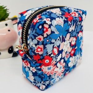 May include: A small, square-shaped zippered pouch with a floral print. The pouch is made of a blue fabric with red, white and pink flowers. The zipper is black with a gold pull.