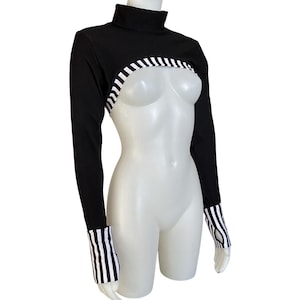 May include: Black long-sleeved crop top with a high neckline. The top features a white and black striped design across the chest and on the cuffs. The garment is displayed on a white mannequin.