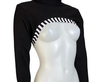Op de afbeelding: Een zwart bolero-vest met lange mouwen en een opstaande kraag, voorzien van een kort model met wit-zwarte verticale strepen langs de onderkant en bij de mouwboorden, inclusief duimgaten in de mouwen.