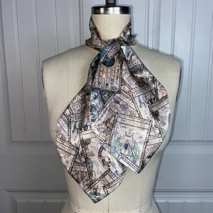 Op de afbeelding: Een beige zijden sjaal met een bedrukt ontwerp van tarotkaarten. De kaarten zijn in verschillende kleuren, waaronder blauw, groen en bruin. De sjaal is om de nek van een mannequin geknoopt.