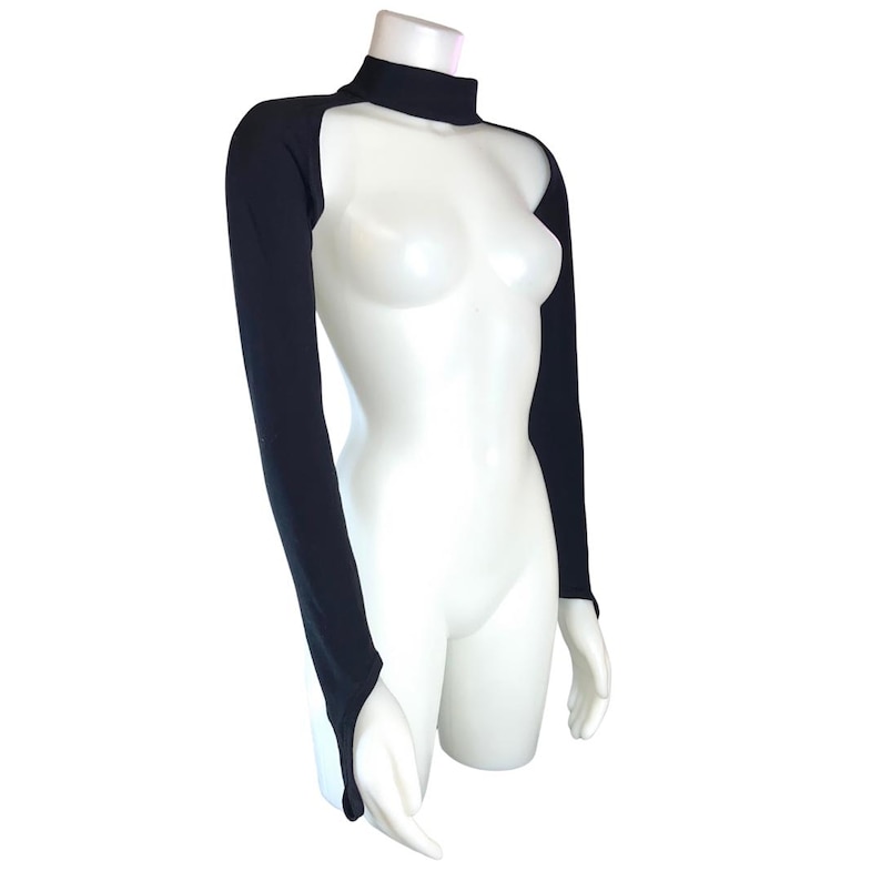 Op de afbeelding: Zwarte shrug met lange mouwen en een hoge halslijn. De shrug heeft duimgaten en wordt getoond op een witte mannequin. Het kledingstuk is gemaakt van een rekbaar materiaal en is ontworpen om de armen en schouders te bedekken.