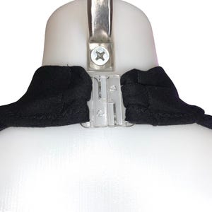 Op de afbeelding: Close-up van een zwart kledingstuk met een doorzichtige plastic sluiting en een metalen hanger. Het kledingstuk is over een witte vorm gedrapeerd, waarbij de halslijn en het sluitmechanisme worden benadrukt.