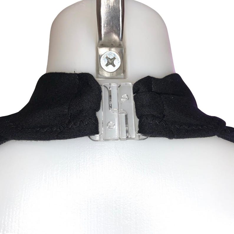 Op de afbeelding: Close-up van een zwart kledingstuk met een doorzichtige plastic sluiting en een metalen hanger. Het kledingstuk is over een witte vorm gedrapeerd, waarbij de halslijn en het sluitmechanisme worden benadrukt.