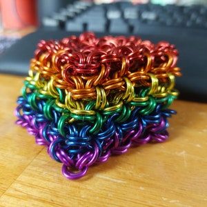May include: A rainbow-colored chainmail heart-shaped sculpture made of metal rings. The sculpture is made of six layers of rings, each layer a different color of the rainbow.