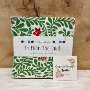 May include: A stack of fabric squares with a green and white botanical pattern, wrapped with a label that reads "IN FROM THE COLD" by Kate Spain for Moda. A small card with the text "Cottonflower Quilts handmade" is also present.