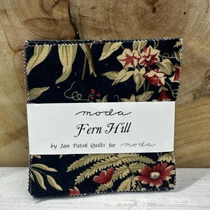 May include: A stack of fabric squares with a floral pattern in navy, red, and beige. The fabric is wrapped with a white label that reads "moda Fern Hill by Jan Patek Quilts for moda."