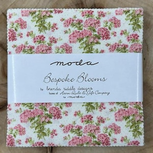 May include: A stack of fabric squares with a floral pattern in pink and green on a cream background. The fabric is wrapped with a white band that reads "moda Bespoke Blooms" by brenda riddle designs.