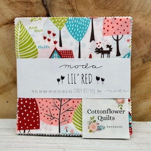 May include: A stack of fabric squares with a whimsical forest print featuring trees, houses, and a wolf. The fabric is wrapped with a label that reads "moda LIL' RED" and "Cottonflower Quilts handmade."