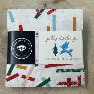 May include: A pack of 42 5-inch charm squares featuring a white background with colourful, abstract, geometric shapes. The label reads "jolly darlings" with a blue reindeer illustration and the text "COLLABORATIVE COLLECTION".