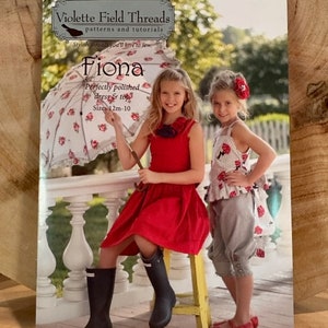 May include: A sewing pattern booklet for a dress and top called "Fiona" by Violette Field Threads. The pattern is for sizes 12 months to 10 years. The cover features two girls wearing the dress and top, holding a white parasol with red flowers.