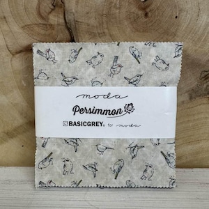 May include: A stack of fabric squares with a light beige background and a repeating pattern of small, black bird illustrations. The fabric is wrapped with a white label that reads "moda Persimmon" and "BASICGREY for moda."