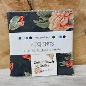 May include: A stack of fabric squares with a floral pattern in shades of pink, green, and gray. The fabric is labeled "ETCHINGS" and "Cottonflower Quilts". The background is a wooden surface.