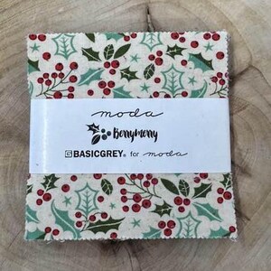 May include: A cream-colored fabric square with a repeating pattern of green holly leaves and red berries. The fabric has a white label with the text "moda Berrymerry BASICGREY for moda".