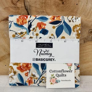 May include: A stack of fabric squares with a floral pattern in orange, blue, and cream. The fabric is labeled "Nutmeg" by BasicGrey. A ruler is placed on the fabric. A small tag reads "Cottonflower Quilts handmade."
