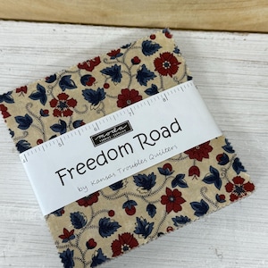 May include: A stack of fabric squares with a floral pattern in red and blue on a tan background. The fabric is wrapped with a white band that reads "Freedom Road" and "by Kansas Troubles Quilters".