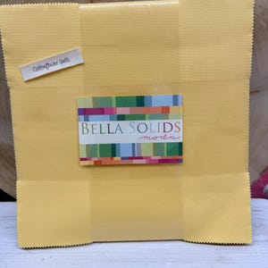 May include: A stack of square yellow fabric pieces wrapped in clear plastic. A rectangular label on the front features the text BELLA SOLIDS moda with a colorful geometric pattern. A small white tag reads Cottonflower Quilts in black ink.