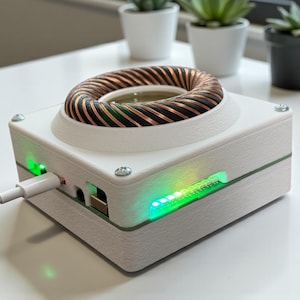 Può includere: Un dispositivo elettronico bianco con una bobina di rame sulla parte superiore e luci verdi. Un cavo di ricarica bianco è collegato. Il dispositivo è su una superficie bianca, con piccole piante in vaso sullo sfondo.