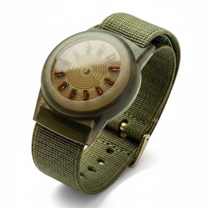 Puede incluir: Un reloj de pulsera de estilo militar con esfera circular y correa de nailon verde oliva. La esfera del reloj tiene una cubierta transparente, un diseño en espiral y marcadores rojos. La caja y la hebilla del reloj también son de color verde oliva.