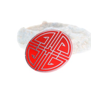 May include: A red circular charm with a silver design of a traditional Chinese knot. The knot is a symbol of good luck and fortune.