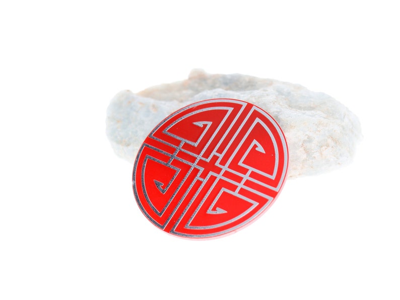 May include: A red circular charm with a silver design of a traditional Chinese knot. The knot is a symbol of good luck and fortune.