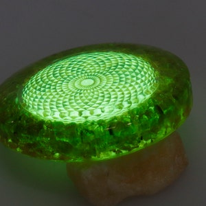 May include: A glowing green, oval-shaped object with a detailed geometric pattern on its surface. The object is made of a translucent material, possibly resin or glass, and rests on a light-colored stone. The intricate design is illuminated from within.