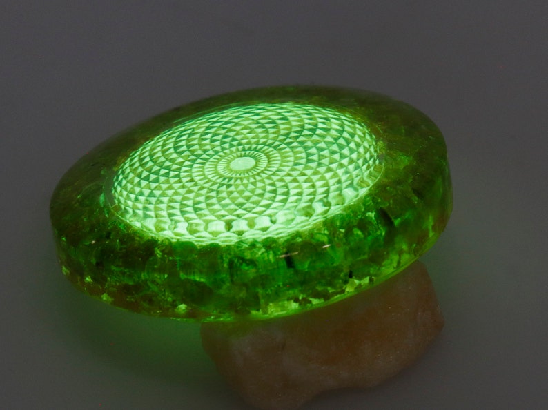 May include: A glowing green, oval-shaped object with a detailed geometric pattern on its surface. The object is made of a translucent material, possibly resin or glass, and rests on a light-colored stone. The intricate design is illuminated from within.