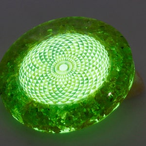 May include: A glowing green resin art piece with a geometric design. The central area features a detailed pattern of triangles and circles, emitting a bright, internal light. The outer edges have a textured, organic appearance.