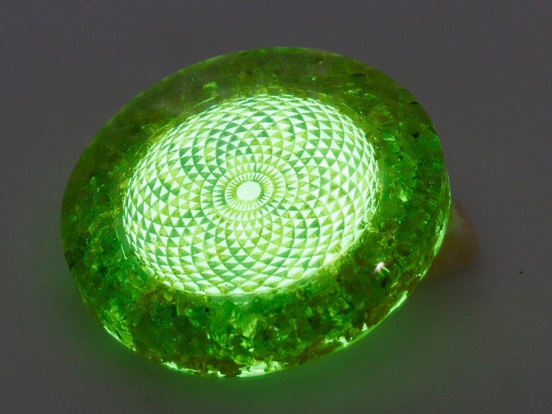 May include: A glowing green resin art piece with a geometric design. The central area features a detailed pattern of triangles and circles, emitting a bright, internal light. The outer edges have a textured, organic appearance.