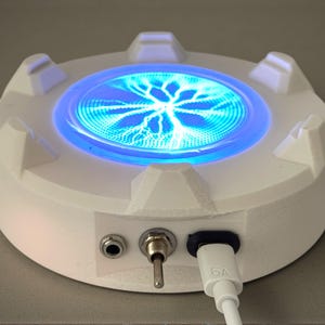 Può includere: Un dispositivo elettronico bianco e rotondo con un centro blu brillante che mostra un motivo a forma di fulmine. Ha un interruttore a levetta, una porta e un cavo di ricarica USB-C. Il dispositivo ha un design futuristico.