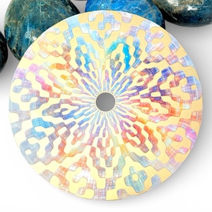 Può includere: Un oggetto decorativo rotondo, color oro, con un foro centrale e un design colorato e fantasia nei toni del blu, rosso e giallo. Circondato da pietre blu e grigie.