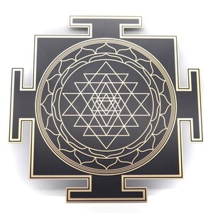 May include: A black and gold Sri Yantra, a geometric design used in Hinduism and Buddhism. The design features interlocking triangles and circles, with a gold outline on a black square. The corners have gold accents.