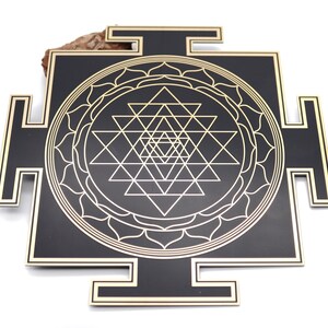 May include: A black and gold Sri Yantra, a geometric design with interlocking triangles and a lotus flower border. The design is set against a black background with gold accents. The object is a square shape with protruding rectangular elements.