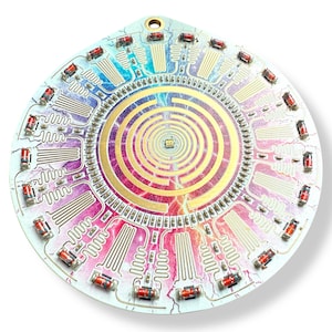 May include: A circular circuit board with a colorful, rainbow-like design. The board features a central spiral pattern with a gold-colored maze. The board is surrounded by a ring of red LED lights.