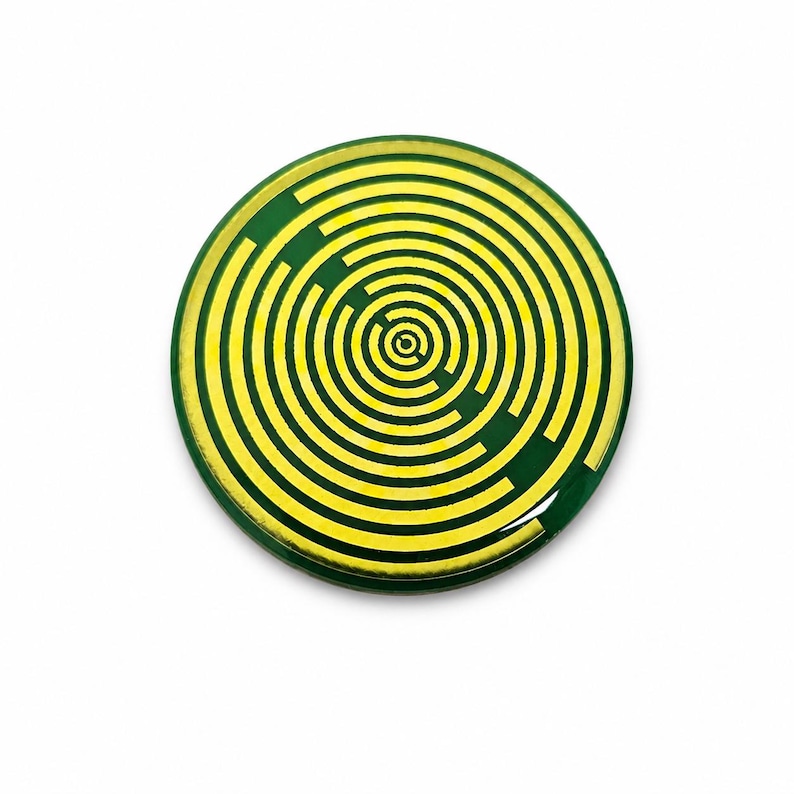 May include: A circular pinback button featuring a dark green background with a bright yellow graphic design consisting of concentric circles and broken line segments that form a maze-like pattern across the entire surface of the round object.