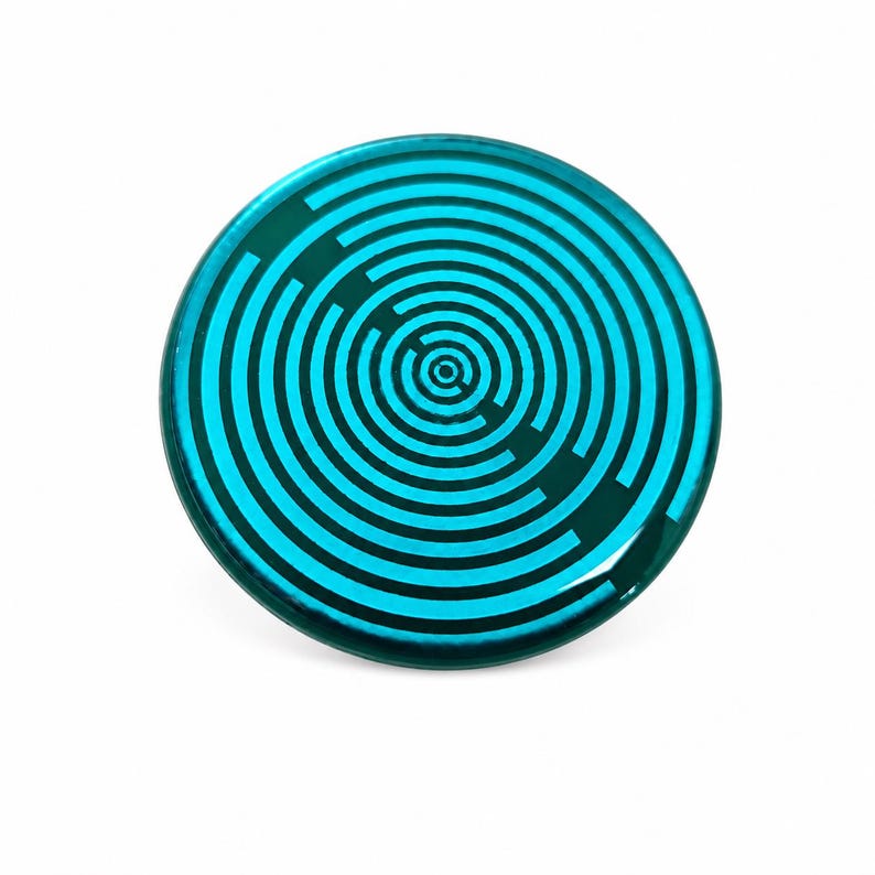May include: A circular button featuring a vibrant cyan and teal colour palette. The surface displays a series of concentric rings that form a maze-like pattern, with distinct breaks in the lines creating a path-like design across the face.