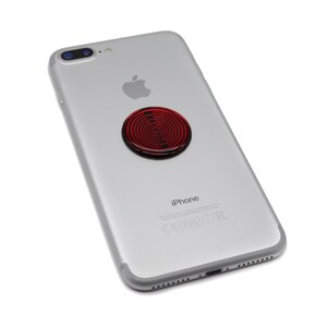 May include: A silver smartphone with a circular red and black spiral pattern accessory attached to the centre of the back panel. The device features a dual camera lens in the upper left corner, an Apple logo, and the text iPhone printed on the back.