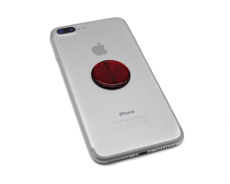 May include: A silver smartphone with a circular red and black spiral pattern accessory attached to the centre of the back panel. The device features a dual camera lens in the upper left corner, an Apple logo, and the text iPhone printed on the back.
