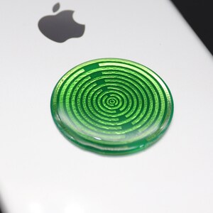 May include: A circular, translucent green adhesive accessory featuring a concentric maze-like pattern of raised lines. The disc is attached to the back of a white smartphone, positioned below the black Apple logo, creating a distinct visual contrast.