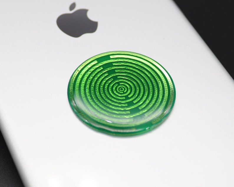 May include: A circular, translucent green adhesive accessory featuring a concentric maze-like pattern of raised lines. The disc is attached to the back of a white smartphone, positioned below the black Apple logo, creating a distinct visual contrast.