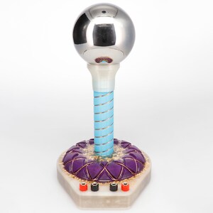 May include: A silver sphere sits atop a blue and white striped column, which is mounted on a hexagonal base with a purple and gold design. Three black and red electrical terminals are visible on the base.